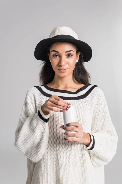 Menina americana africana atraente em elegante suéter branco e chapéu segurando lata branca e olhando para a câmera isolada no branco — Fotografia de Stock