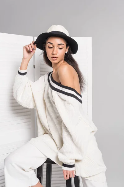 Atractiva chica afroamericana en ropa blanca con estilo y sombrero posando cerca de la pantalla plegable en blanco - foto de stock