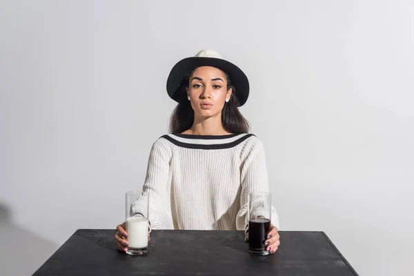 Atractiva chica afroamericana en ropa blanca con estilo y sombrero sentado en la mesa negra con leche y refrescos en vasos en blanco - foto de stock