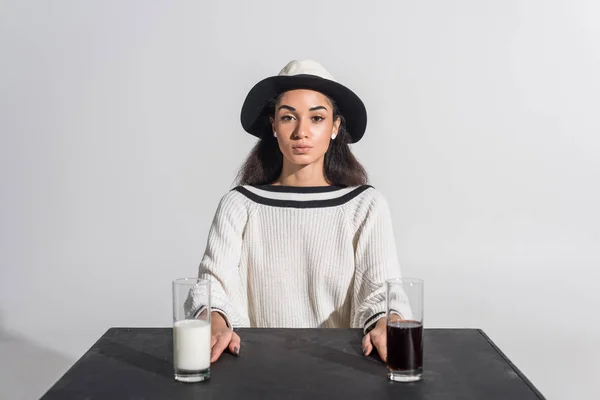 Beautiful african american woman in stylish white clothes and hat sitting at black table with milk and soda in glasses on white — Stock Photo