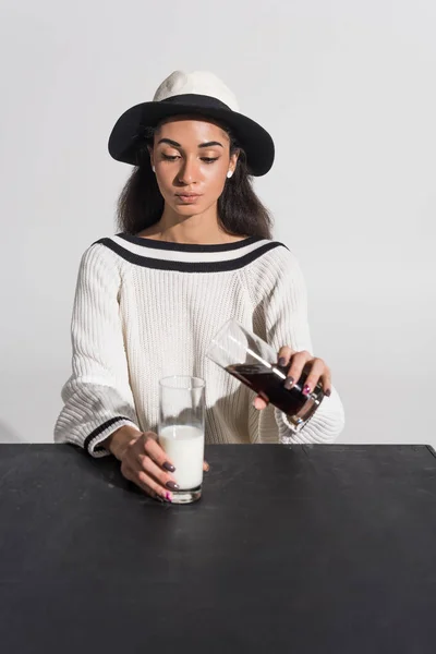 Atractiva chica afroamericana en ropa blanca con estilo y sombrero vertiendo soda en la leche sobre blanco - foto de stock