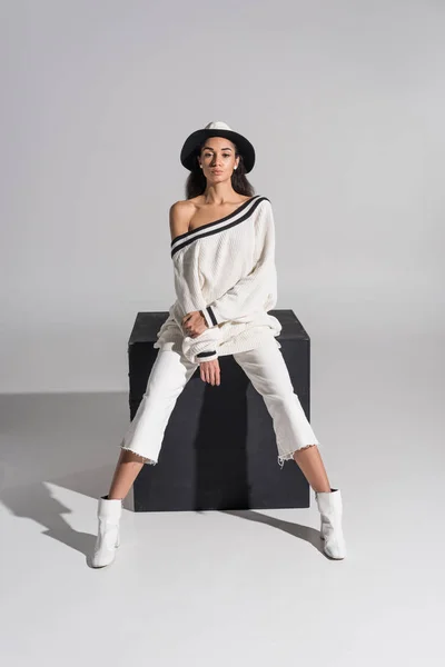 Atractiva chica afroamericana en ropa blanca con estilo y sombrero sentado en cubo negro y mirando a la cámara en blanco - foto de stock