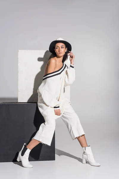 Atractiva chica afroamericana en ropa blanca con estilo y sombrero sentado en cubo negro y mirando hacia otro lado en blanco - foto de stock