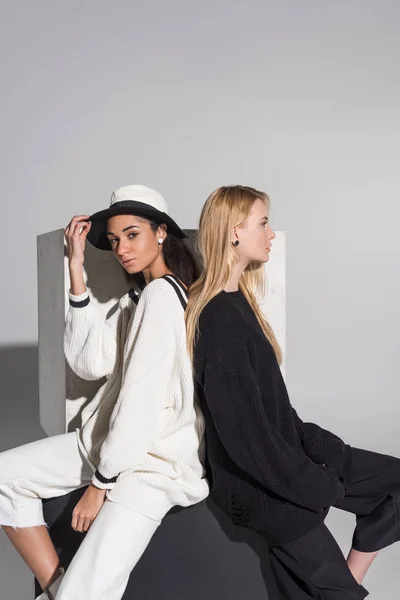 Side view of beautiful multicultural women in black and white clothes sitting back to back on white — Stock Photo