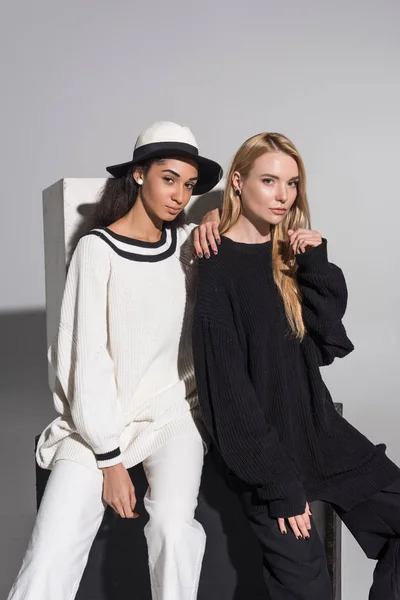Schöne multikulturelle Frauen in schwarz-weißer Kleidung, die auf einem schwarzen Würfel sitzen und in die Kamera in weißer Kleidung schauen — Stockfoto