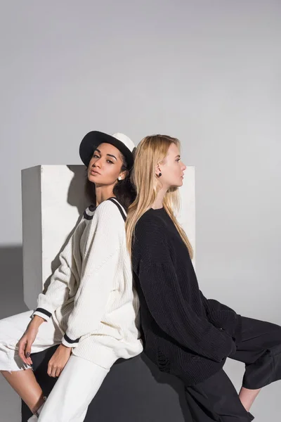 Side view of attractive multicultural women in black and white clothes sitting back to back on white — Stock Photo