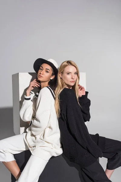 Side view of attractive multicultural women in black and white clothes sitting back to back and looking at camera on white — Stock Photo