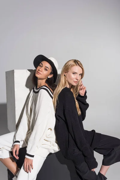 Side view of attractive multicultural women in stylish black and white clothes sitting back to back on white — Stock Photo