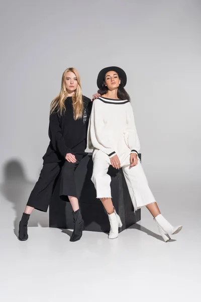 Hermosas mujeres multiétnicas en ropa blanca y negra sentadas en cubo negro y mirando a la cámara en blanco - foto de stock