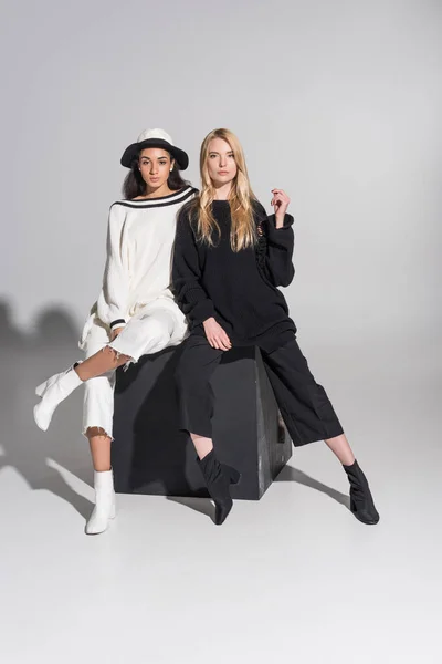 Bellissimi modelli femminili multiculturali in abiti alla moda in bianco e nero seduti sul cubo nero e guardando la fotocamera su bianco — Foto stock