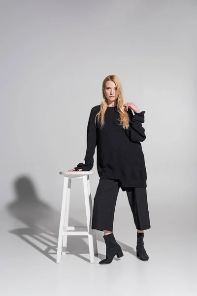 Attractive caucasian blonde woman in stylish black clothes leaning on chair on white — Stock Photo