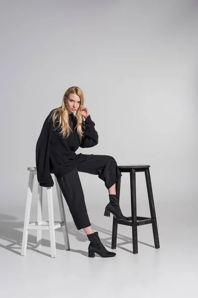 Bela mulher loira caucasiana em roupas pretas sentado em cadeiras e olhando para a câmera no branco — Fotografia de Stock