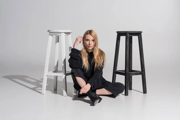 Attractive blonde woman in stylish black clothes sitting near chairs and looking at camera on white — Stock Photo