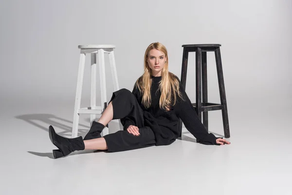 Schöne kaukasische blonde Frau in schwarzen Kleidern sitzt in der Nähe von Stühlen und schaut in die Kamera auf weiß — Stockfoto