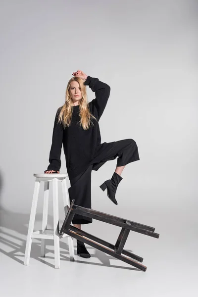 Beautiful caucasian blonde woman in black clothes posing and leaning on chair on white — Stock Photo