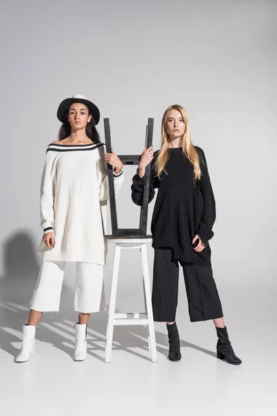 Beautiful multicultural women in black and white clothes standing near chairs on white — Stock Photo