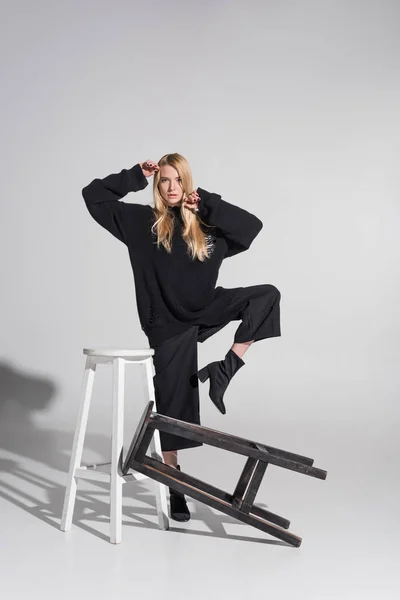 Beautiful caucasian blonde woman in black clothes posing near chairs on white — Stock Photo