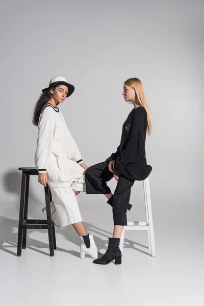 Side view of beautiful multiethnic women in black and white clothes sitting on chairs on white — Stock Photo