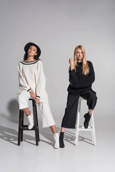 Belle donne multiculturali in abiti alla moda in bianco e nero seduti su sedie e guardando la fotocamera su bianco — Foto stock