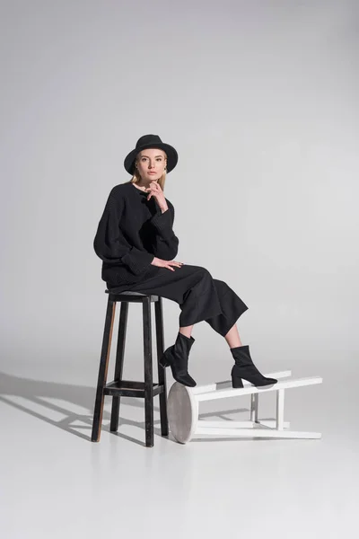 Schöne kaukasische blonde Frau in schwarzer Kleidung und Hut sitzt auf Stuhl und blickt in die Kamera auf weiß — Stockfoto