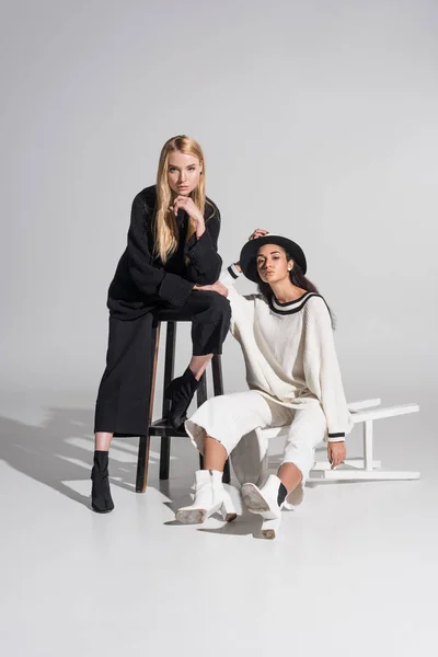 Attraktive multiethnische Frauen in schwarz-weißer Kleidung posieren auf weißen Stühlen — Stockfoto