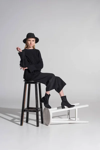 Beautiful caucasian blonde model in stylish black clothes and hat sitting on chair on white — Stock Photo