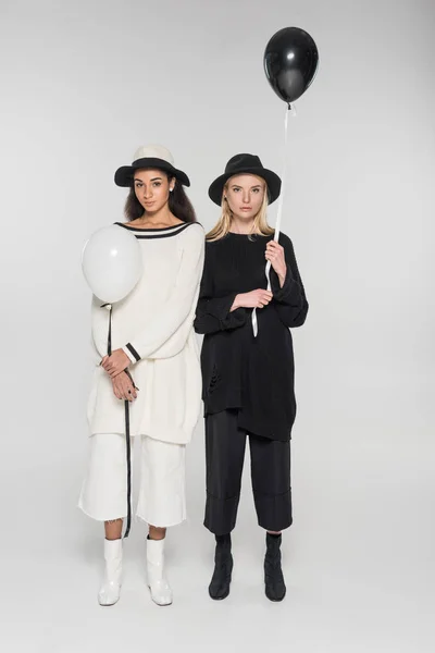 Hermosas mujeres multiculturales en ropa blanca y negra sosteniendo dos globos y mirando a la cámara en blanco - foto de stock