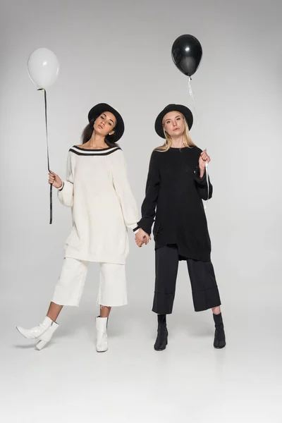 Beautiful multicultural lesbian couple in black and white clothes holding hands and two balloons on white, looking at camera — Stock Photo