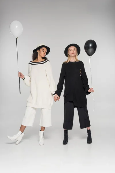 Hermosa pareja de lesbianas multiétnicas en ropa blanca y negra cogidas de la mano y dos globos en blanco - foto de stock