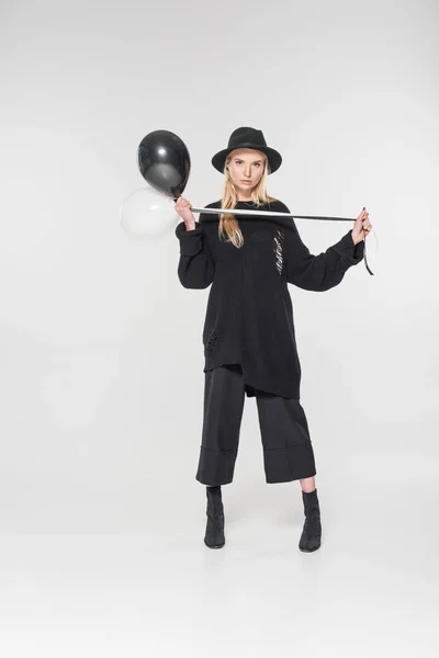 Beautiful caucasian blonde model in black clothes and hat holding balloons on white, looking at camera — Stock Photo