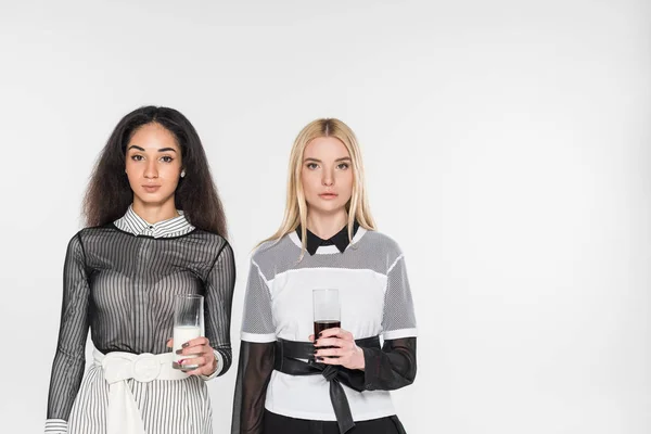 Mulheres multiculturais bonitas em roupas pretas e brancas segurando copos de leite e refrigerante isolado no branco — Fotografia de Stock