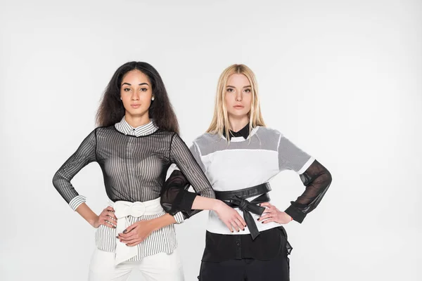 Chicas multiétnicas atractivas en ropa blanca y negra con manos akimbo mirando a la cámara aislada en blanco - foto de stock