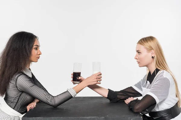 Vista lateral de atractivas chicas multiculturales en ropa blanca y negra sosteniendo vasos con leche y refrescos aislados en blanco - foto de stock