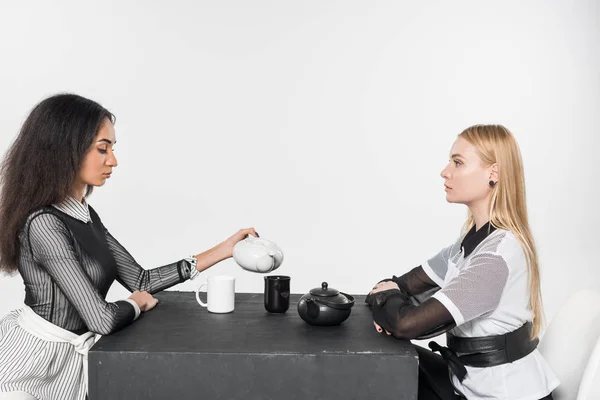 Vista laterale di attraenti ragazze multietniche in bianco e nero vestiti seduti a tavola con teiere e tazze isolate su bianco — Foto stock