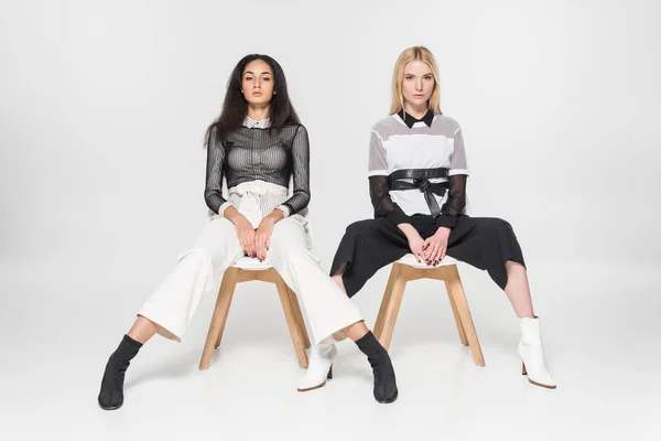 Attractive multiethnic women in black and white clothes posing on chairs and looking at camera isolated on white — Stock Photo