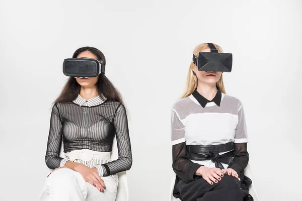 Mulheres multiétnicas atraentes usando fones de ouvido de realidade virtual sentados em cadeiras isoladas em branco — Fotografia de Stock