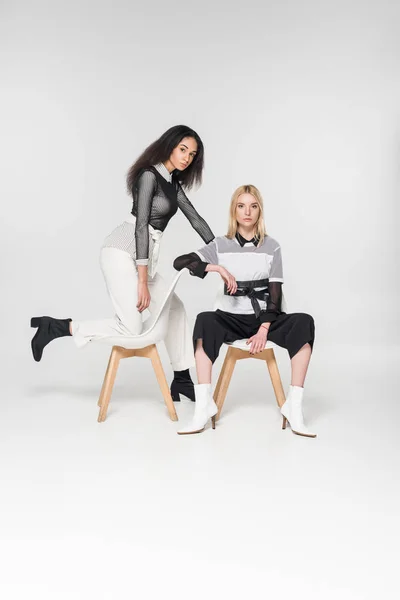 Schöne multikulturelle Mädchen in schwarz-weißer Kleidung, die auf Stühlen sitzen und vereinzelt in die Kamera schauen — Stockfoto