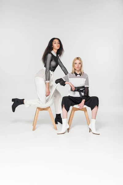 Attraktive multiethnische Frauen in stilvoller schwarzer und weißer Kleidung sitzen auf Stühlen isoliert auf weißen Stühlen — Stockfoto