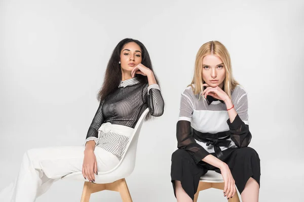 Attractive multiethnic women in trendy black and white clothes sitting on chairs and looking at camera isolated on white — Stock Photo