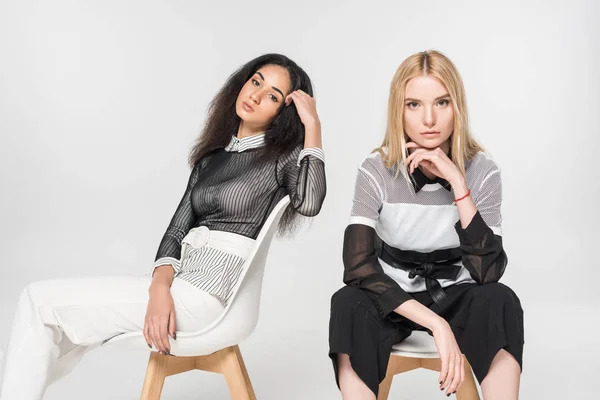 Attractive multiethnic brunette and blonde women in black and white clothes sitting on chairs isolated on white — Stock Photo