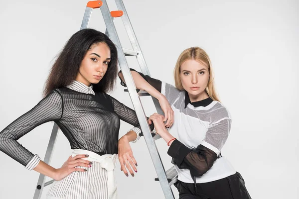 Attractive multiethnic women in black and white clothes leaning on ladder and looking at camera isolated on white — Stock Photo