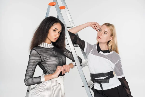 Attractive multiethnic women in black and white clothes leaning on ladder isolated on white — Stock Photo