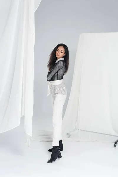 Bella ragazza afro-americana in abiti alla moda in piedi vicino a tulle su bianco e guardando giù — Foto stock