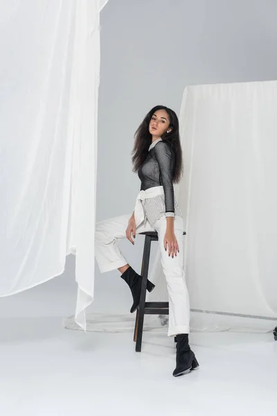 Beautiful african american girl in stylish clothes sitting on chair near tulle on white — Stock Photo