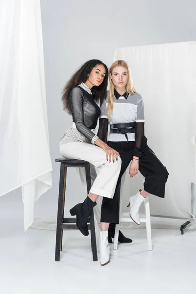 Attractive multiethnic women in black and white clothes sitting on chairs and looking at camera on white — Stock Photo