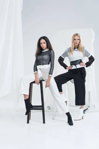 Attractive multiethnic women in trendy black and white clothes posing on chairs on white — Stock Photo