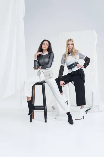 Belle donne multietniche in abiti alla moda in bianco e nero in posa su sedie su bianco — Foto stock