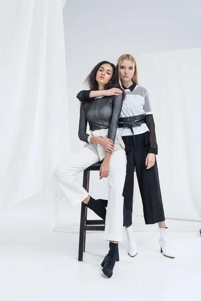 Attractive multiethnic girlfriends in black and white clothes sitting on chairs and hugging on white — Stock Photo