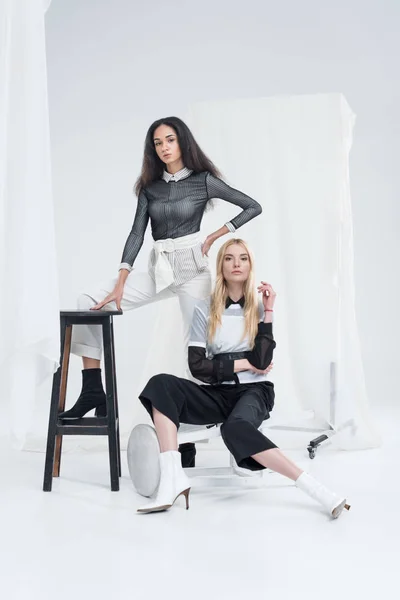 Привабливі багатоетнічні жінки в стильному чорно-білому одязі позує на стільцях біля тюлю на білому — Stock Photo