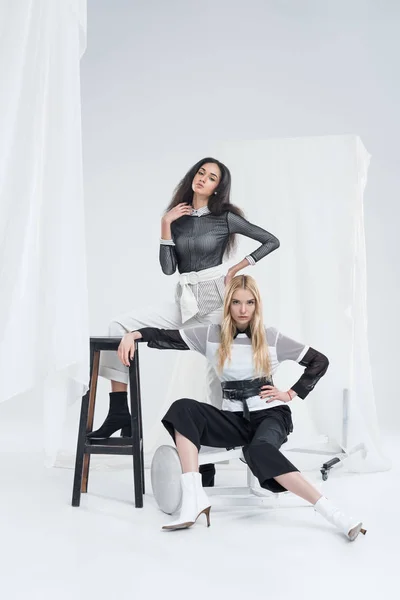 Attractive multicultural women in black and white clothes posing on chairs near tulle on white — Stock Photo